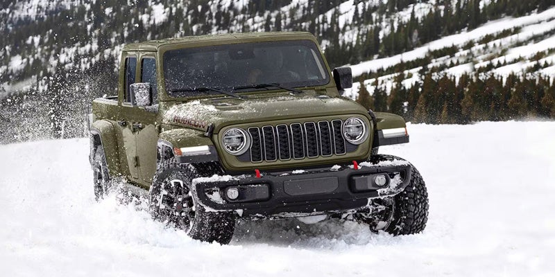 Green Jeep Gladiator driving through snowy terrain in a winter mountain setting.