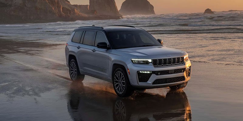 White Jeep Grand Cherokee parked on a beach with ocean and rocky coastline in the background.