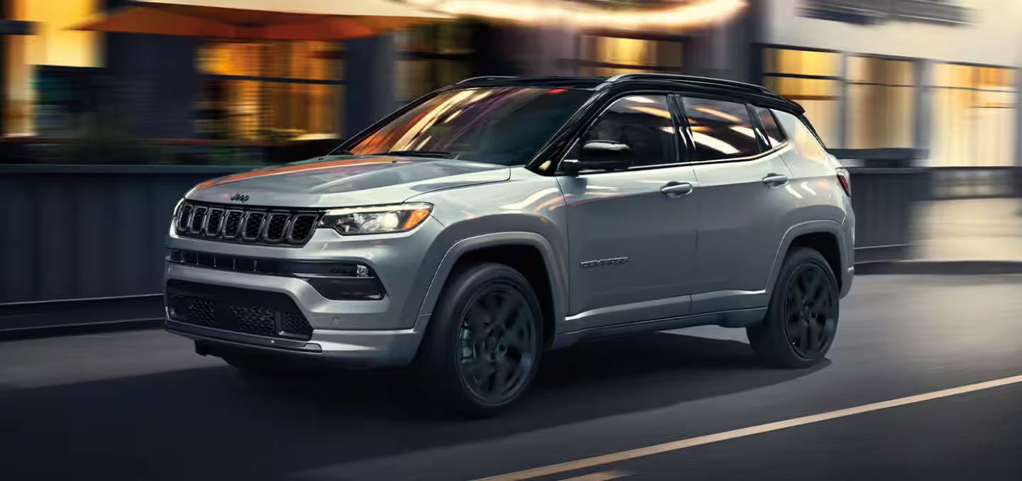 2025 Jeep Compass driving through a modern city street at dusk, showcasing its sleek silver exterior.