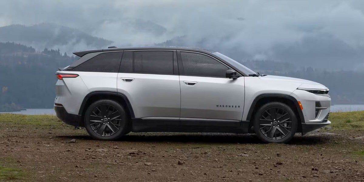 Side view of a silver 2025 Wagoneer S parked on a dirt overlook with misty mountains and a lake in the background, showcasing its sleek electric SUV design.