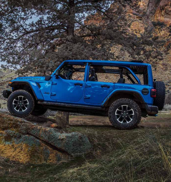 Blue Jeep Wrangler Rubicon climbing over a large rock in a wooded area.