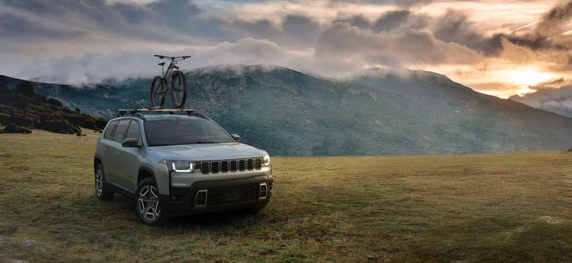 Silver 2026 Jeep Cherokee parked on a grassy field with a bicycle mounted on the roof, mountains and sunset in the background, for Windward Dodge Chrysler Jeep RAM.